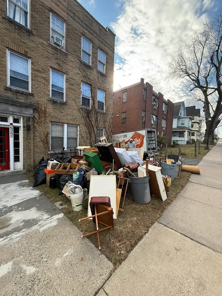 Pile of discarded furniture and debris in front of a brick apartment building on a sunny day.