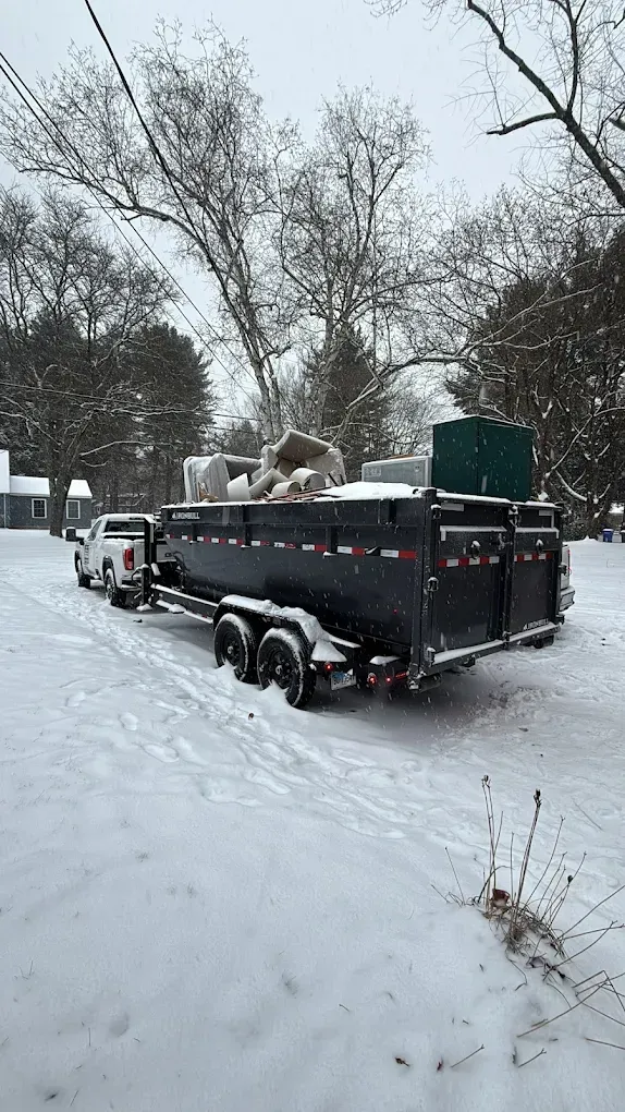 White pickup truck towing a black trailer filled with debris, parked on a snow-covered road.