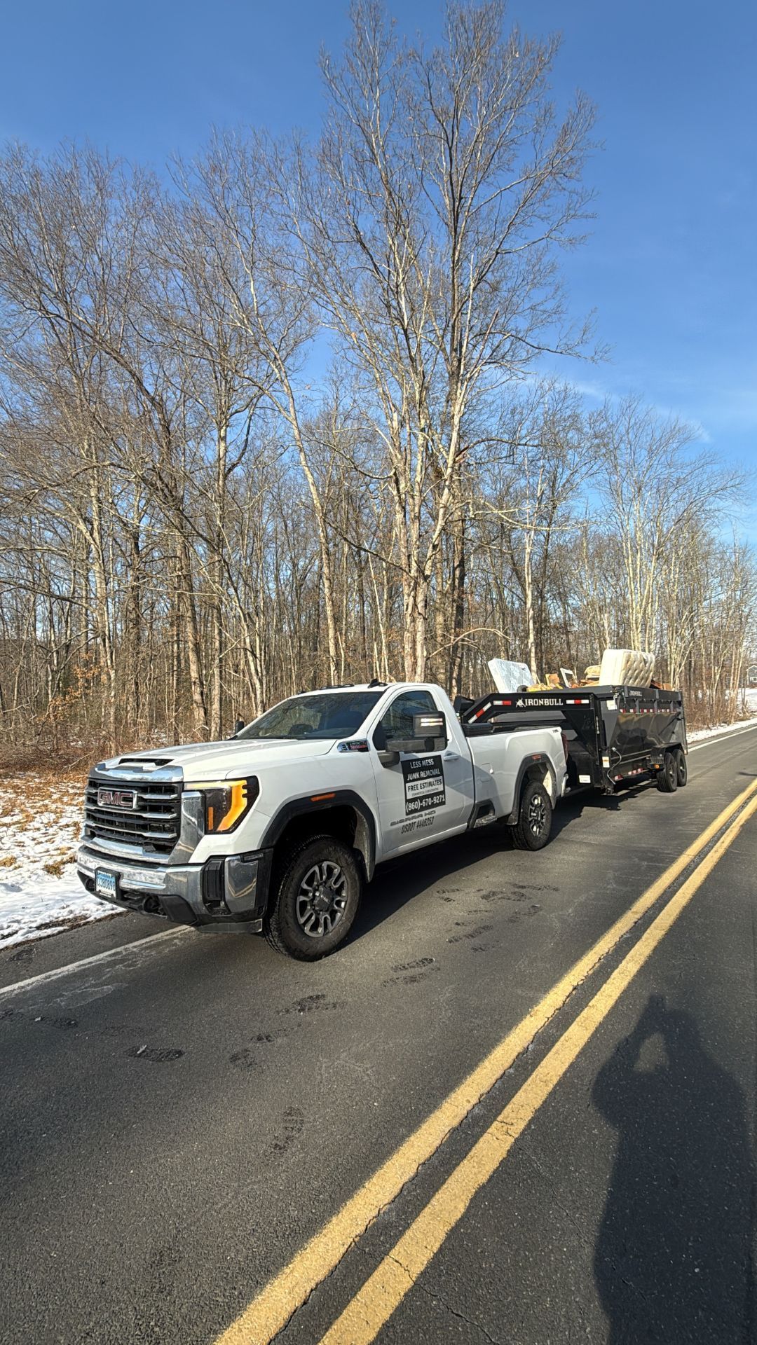 White pickup truck towing a trailer on a road with trees in the background. Sunny, winter setting.