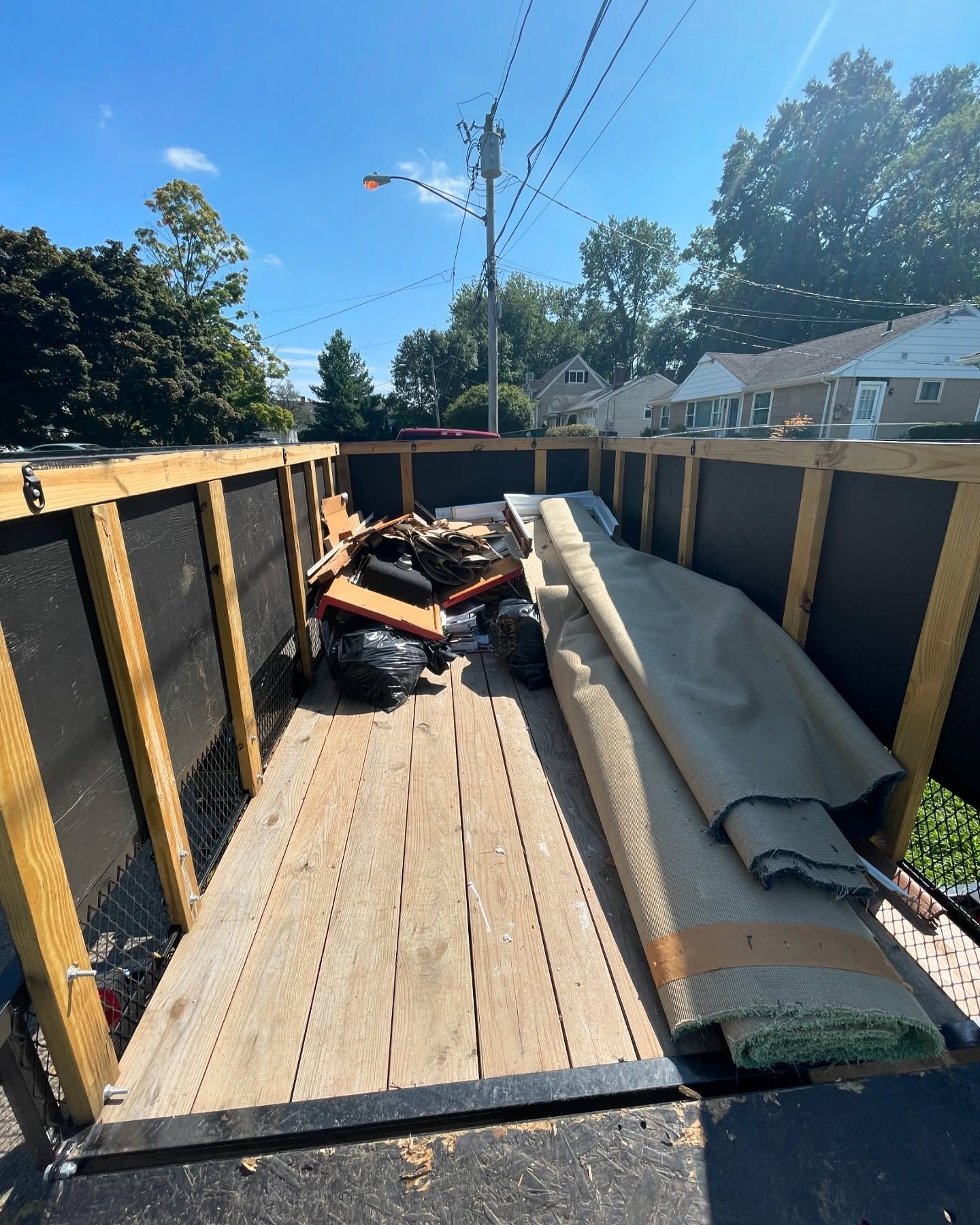 A wooden trailer filled with debris and rolled-up carpet, parked on a sunny street.