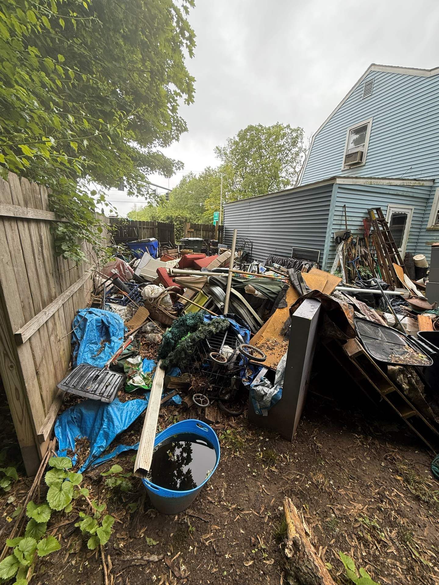Backyard filled with trash and debris; a blue house is visible. Overcast sky.