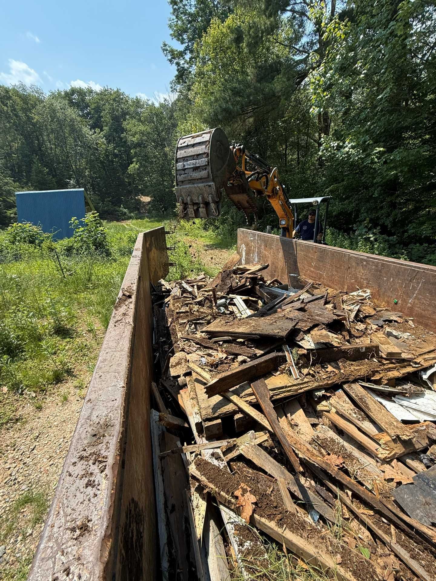 Excavator loading debris into a large metal container in a wooded area.