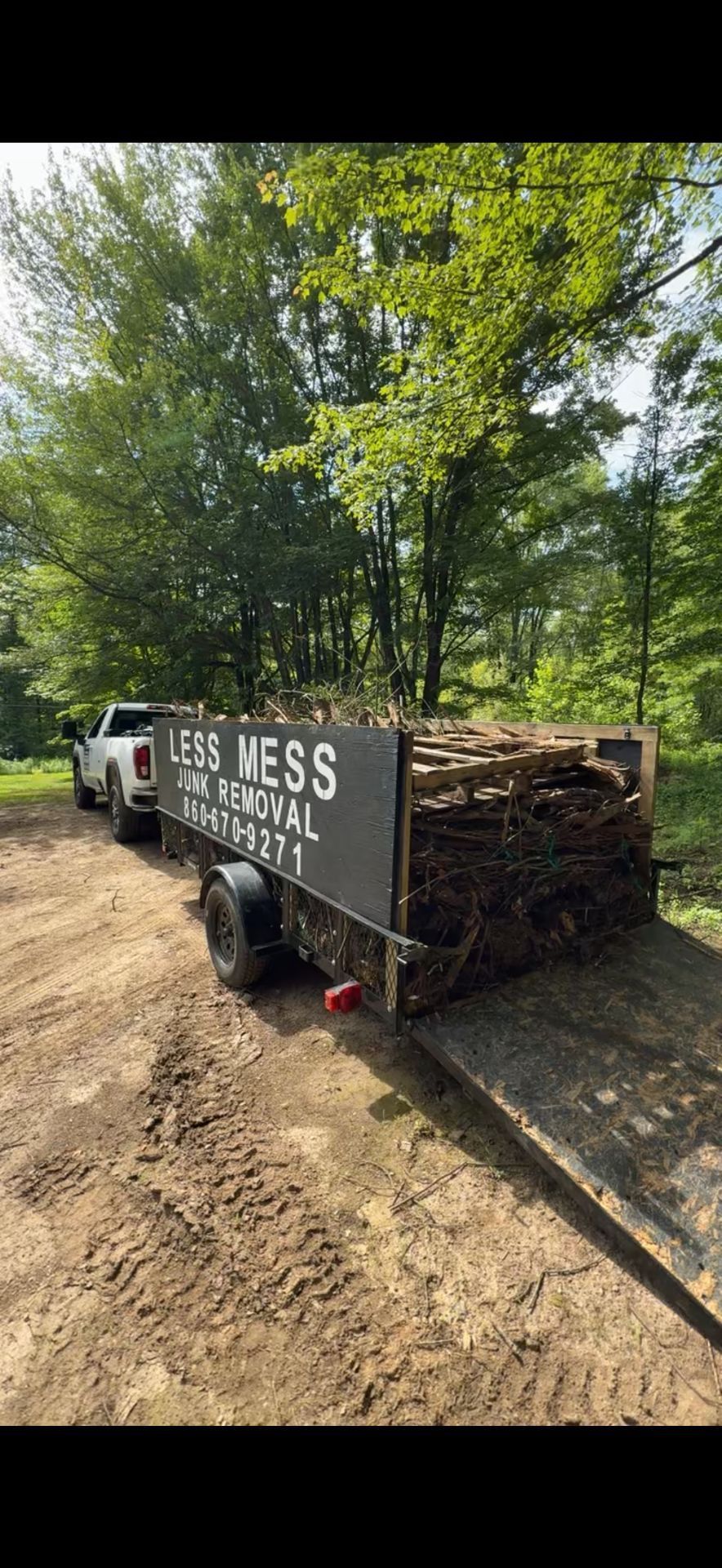 A white truck pulling a trailer loaded with wood debris, in a wooded area. The trailer has 