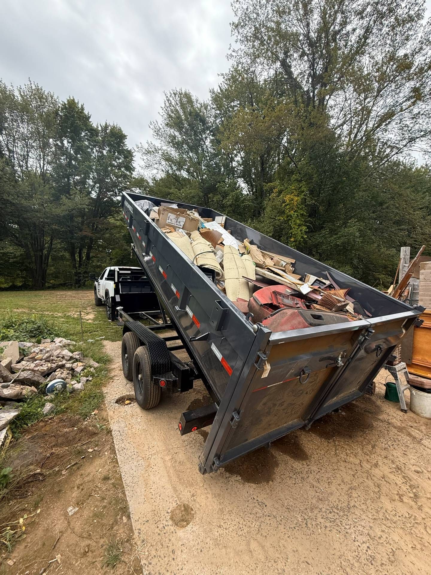 Dump truck with bed raised, full of debris, backed up on a dirt driveway next to a grassy area with trees under an overcast sky.