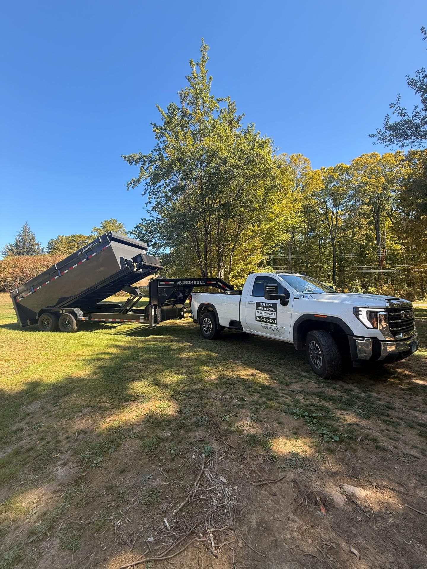 White pickup truck towing a black dump trailer on a grassy field with trees in the background.