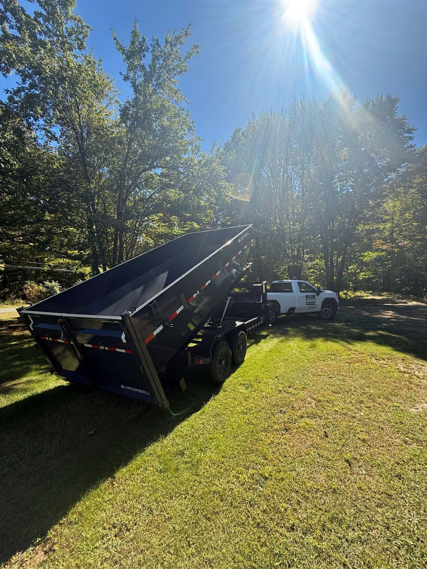 Black dump trailer tilted up, with a white truck. Green grass and trees in sunlight.