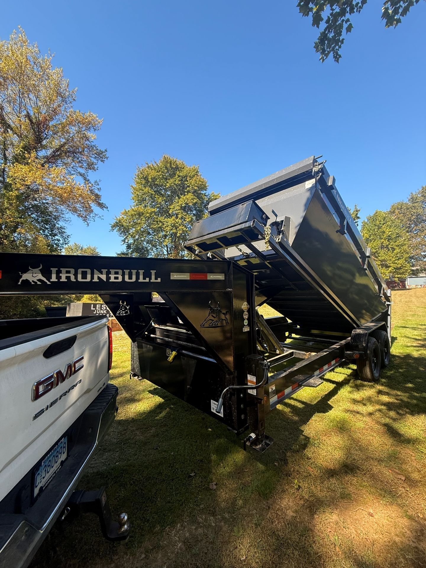Black Ironbull dump trailer attached to a white GMC truck, raised bed, parked on grass, blue sky.