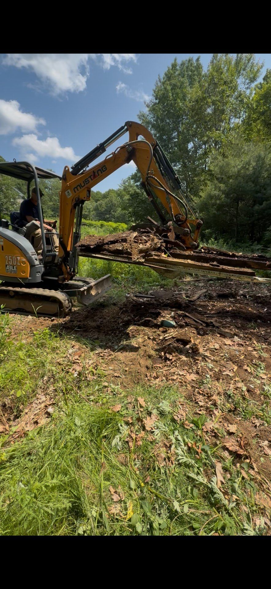 Excavator moving dirt in a grassy field, trees in the background under a blue sky.
