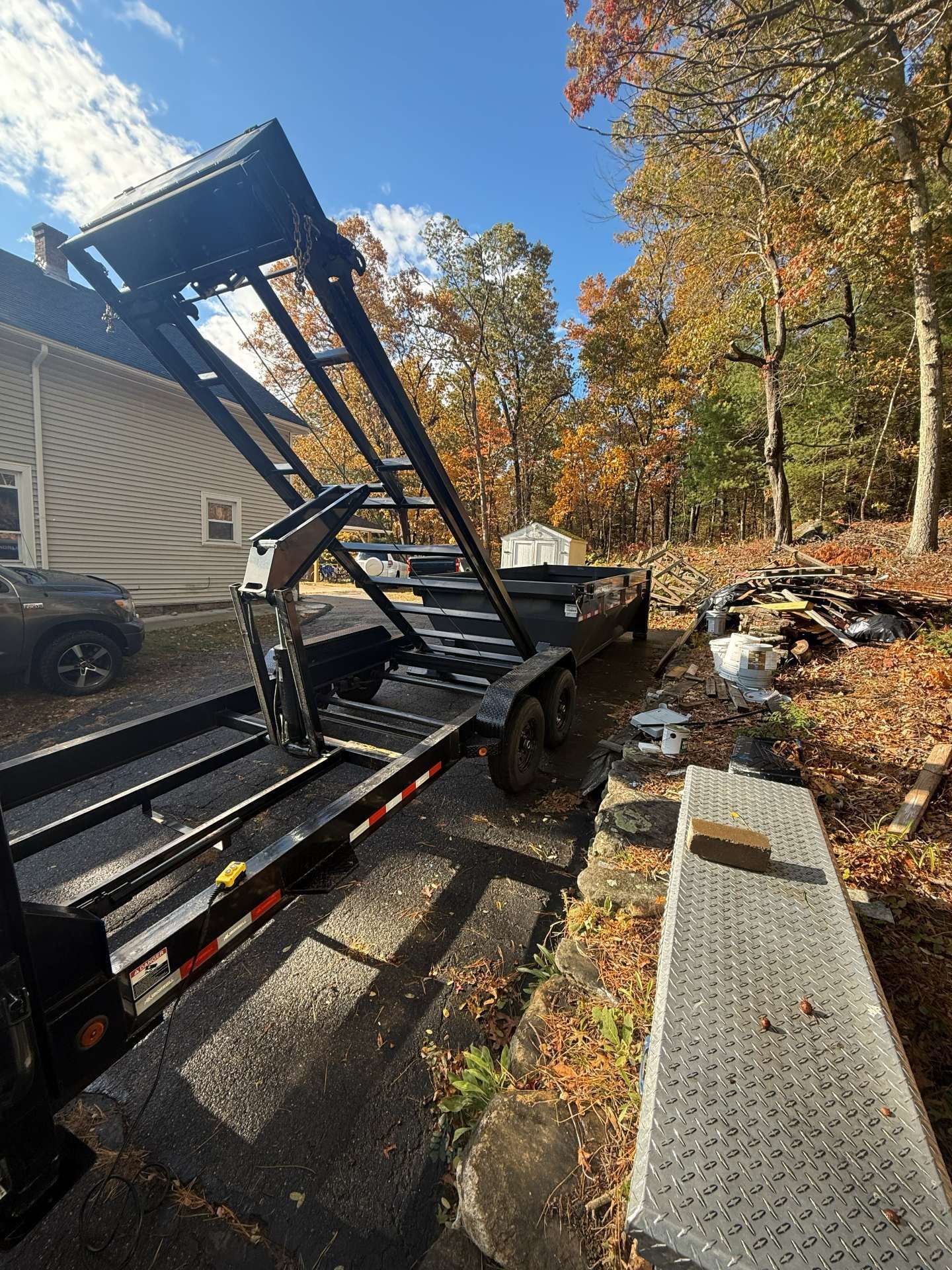 Black dump trailer, tilted, next to a house and foliage, on a sunny day.