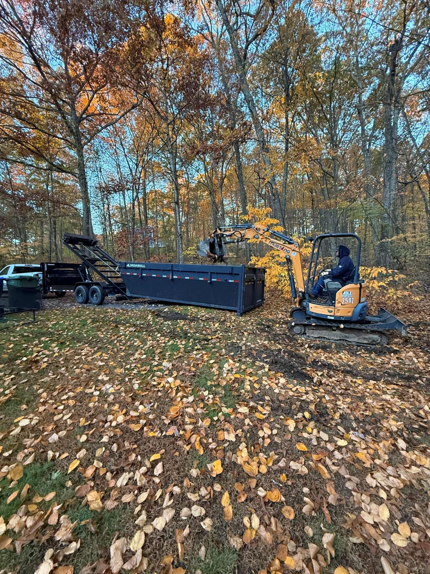 A compact excavator and trailer with a long black box, in a wooded area with fall foliage.