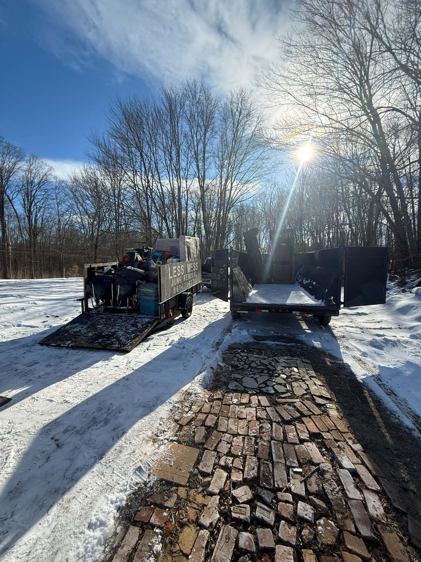 Two trailers on a snowy brick road under a bright sun. Trees are in the background. One trailer is loaded with debris.