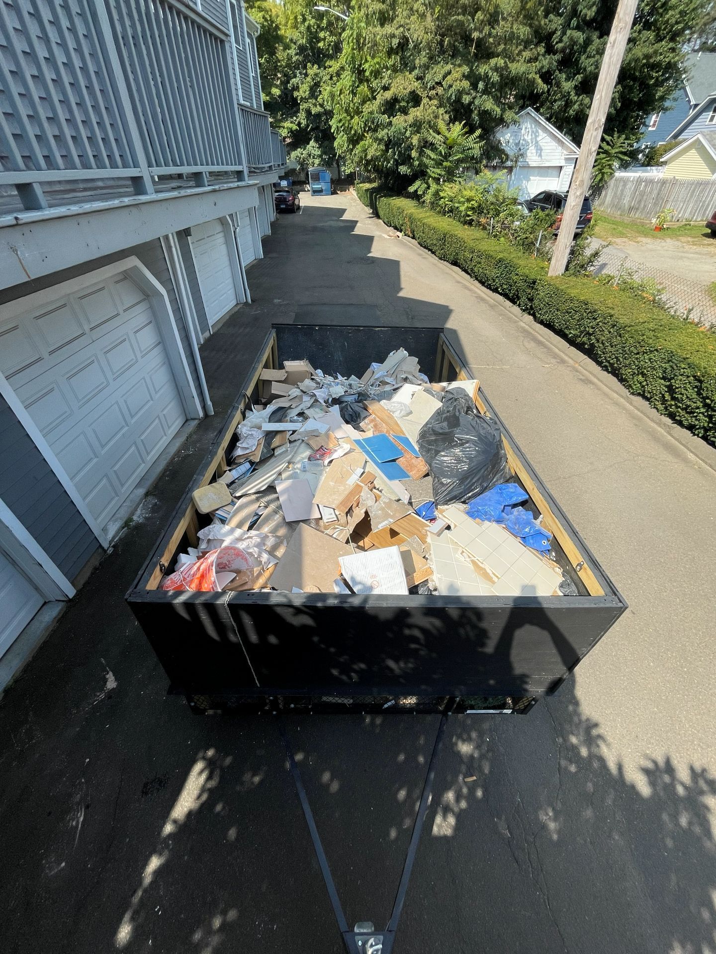 Black dumpster filled with construction debris, parked on a paved driveway next to a building and greenery.