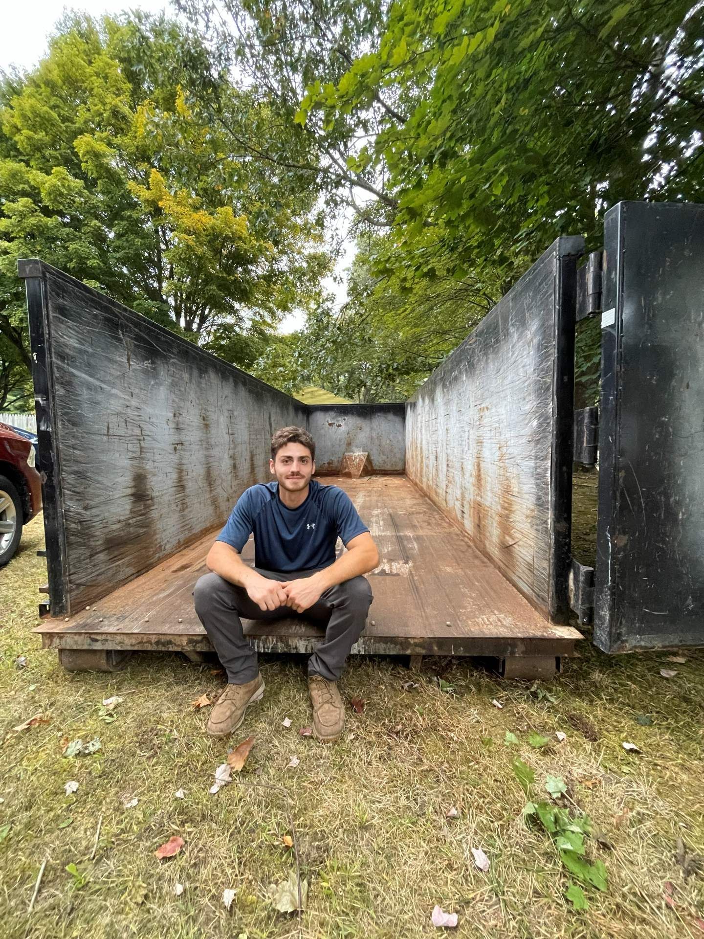Man seated inside a large, open metal container in a grassy area, trees in the background.