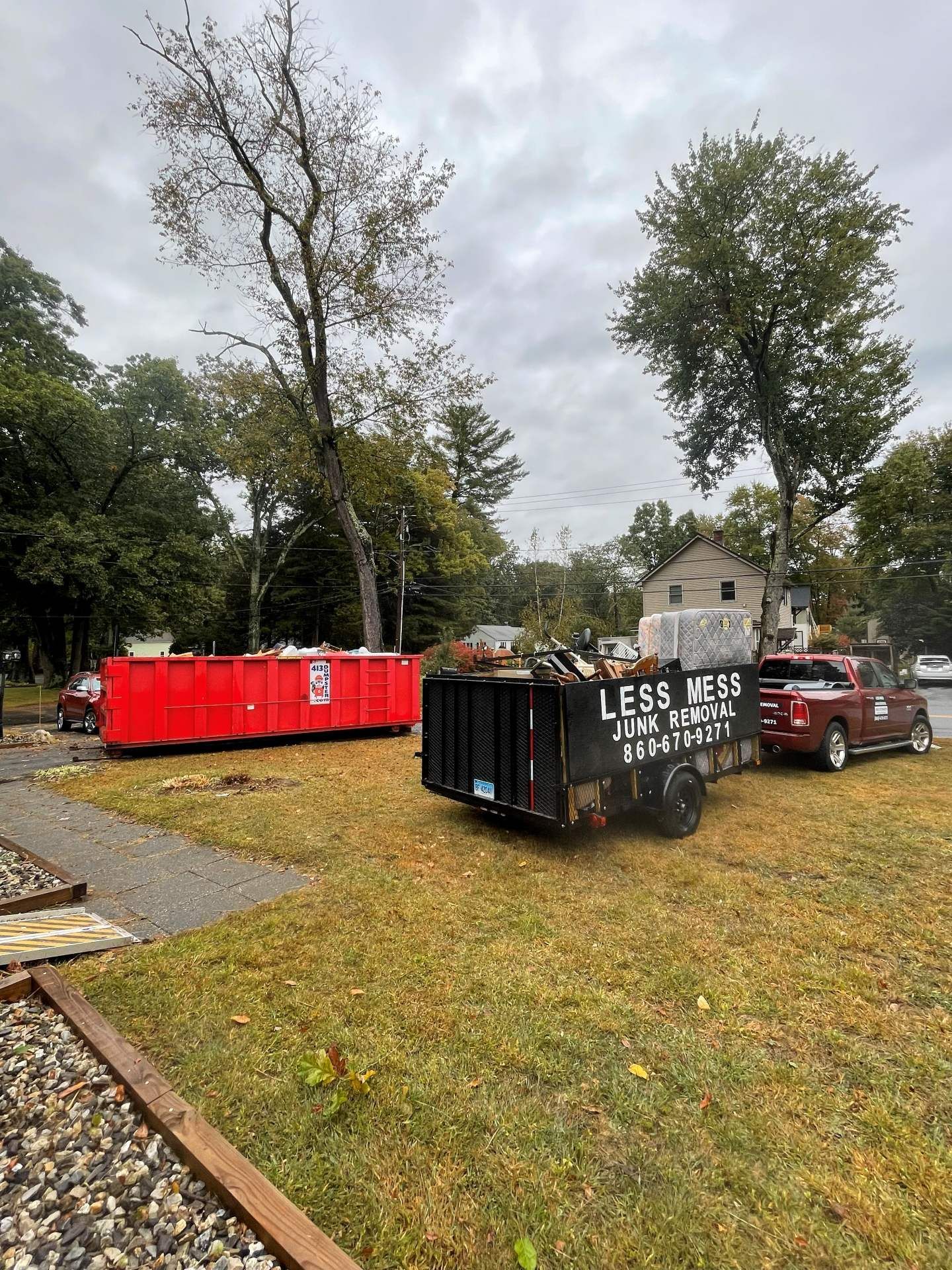 Red dumpster and black trailer filled with debris sit on grass near trees and a building.