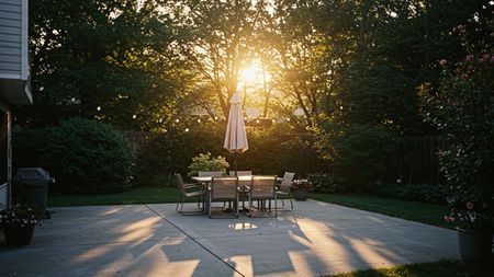 Patio with table and chairs, sun shining through trees.