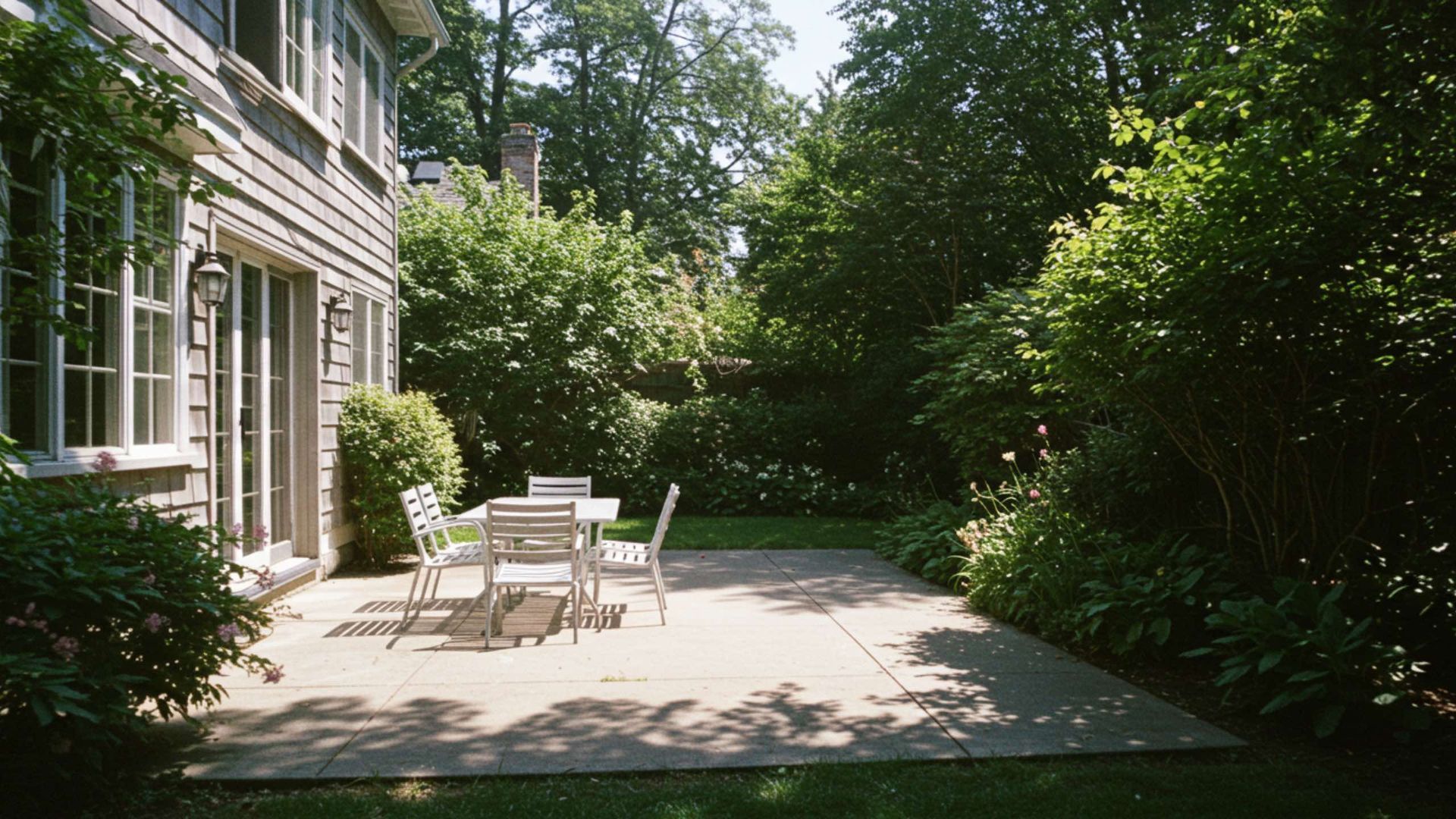 A patio with table and chairs next to a house, surrounded by lush greenery.