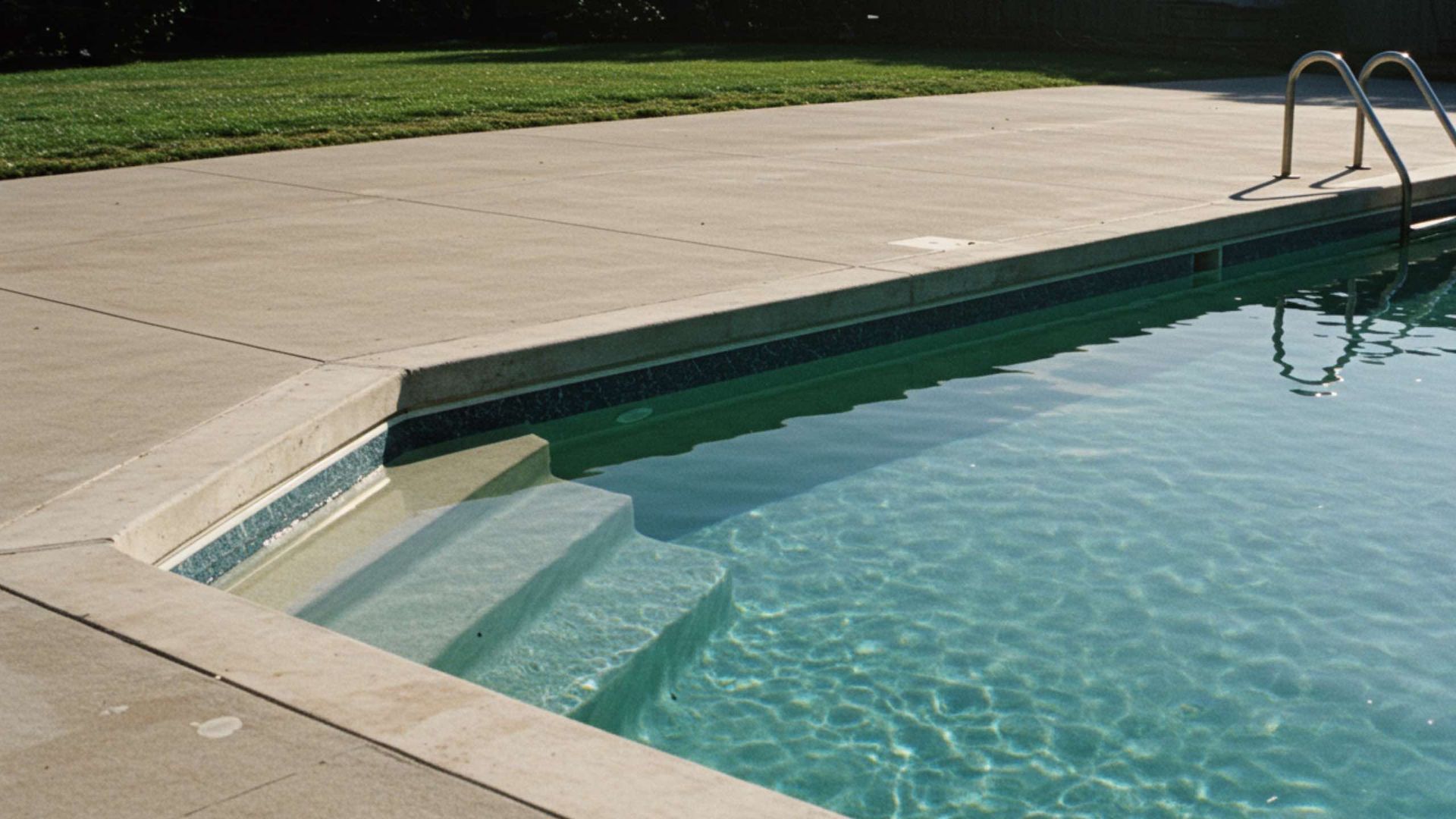A concrete pool deck bordering a swimming pool with steps leading into the shallow end and metal railings in the water.