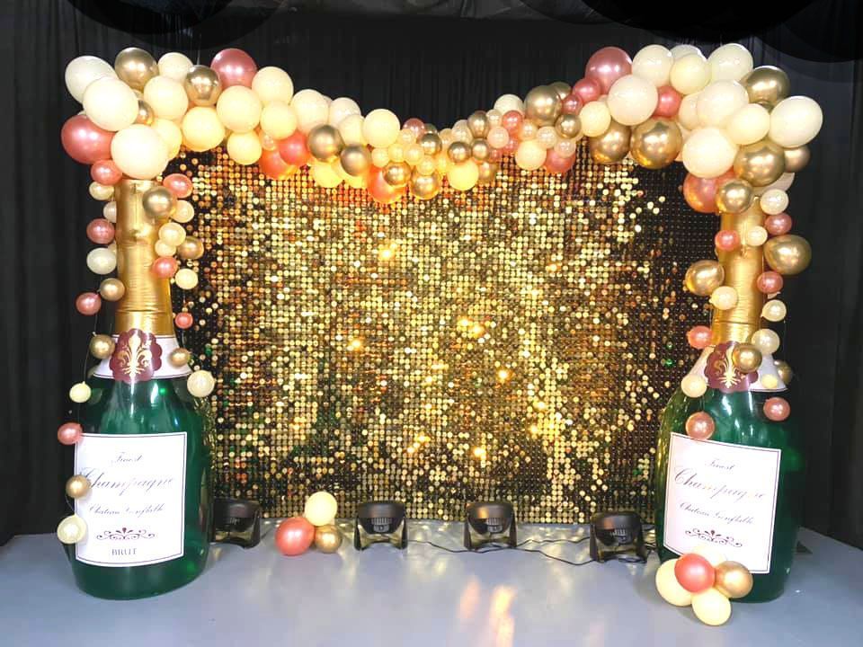Two bottles of champagne are decorated with balloons in front of a gold sequined wall.