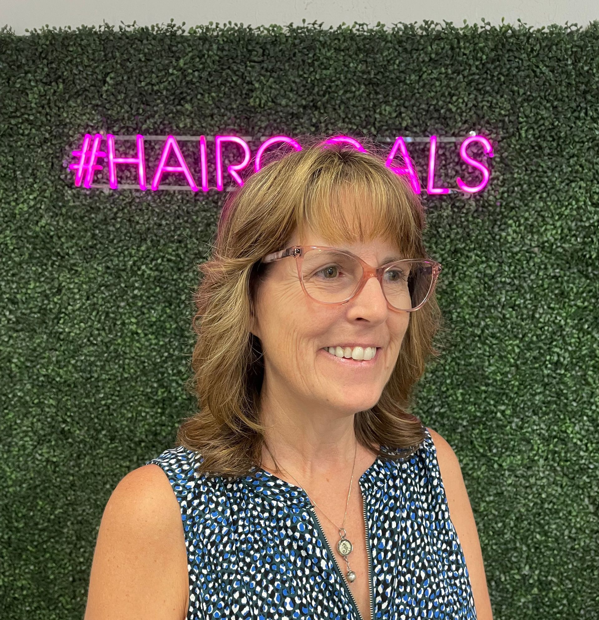 A woman wearing glasses is smiling in front of a neon sign.