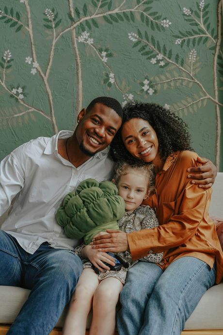 Family of three sitting on a couch, smiling and hugging in front of a green floral wall.