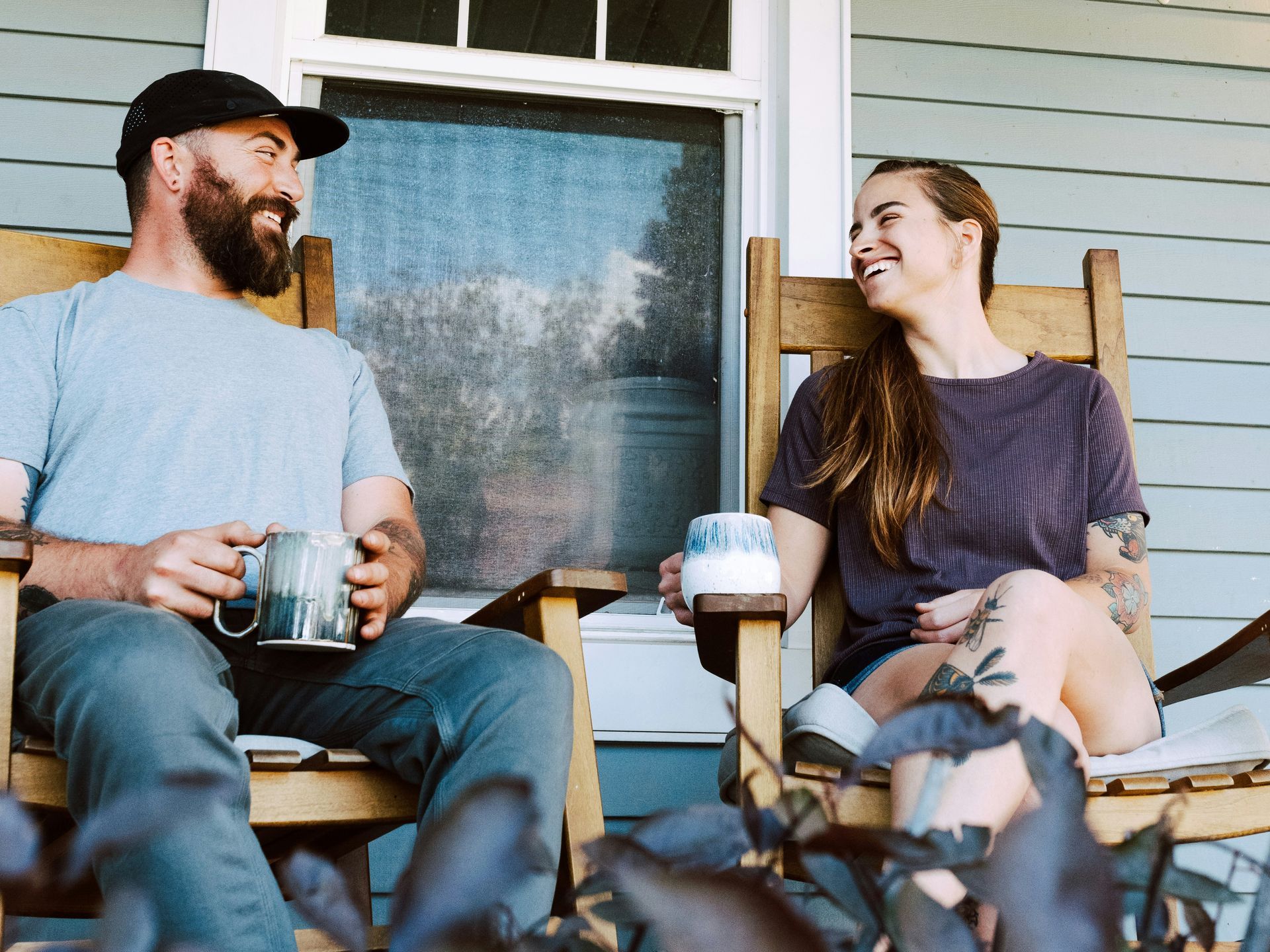 Two people sitting on a porch, smiling and talking over coffee with a dog in the foreground