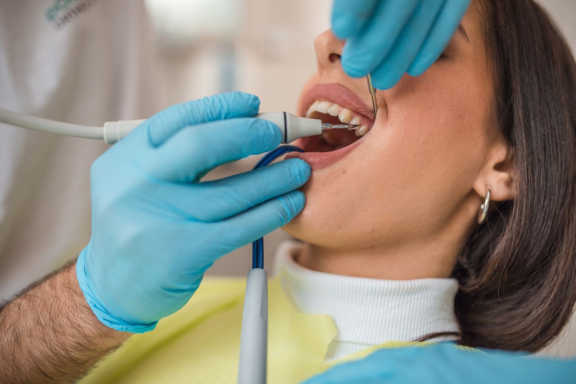 Dentist using dental tools to examine patient's open mouth in a dental office; blue gloves, white coat.