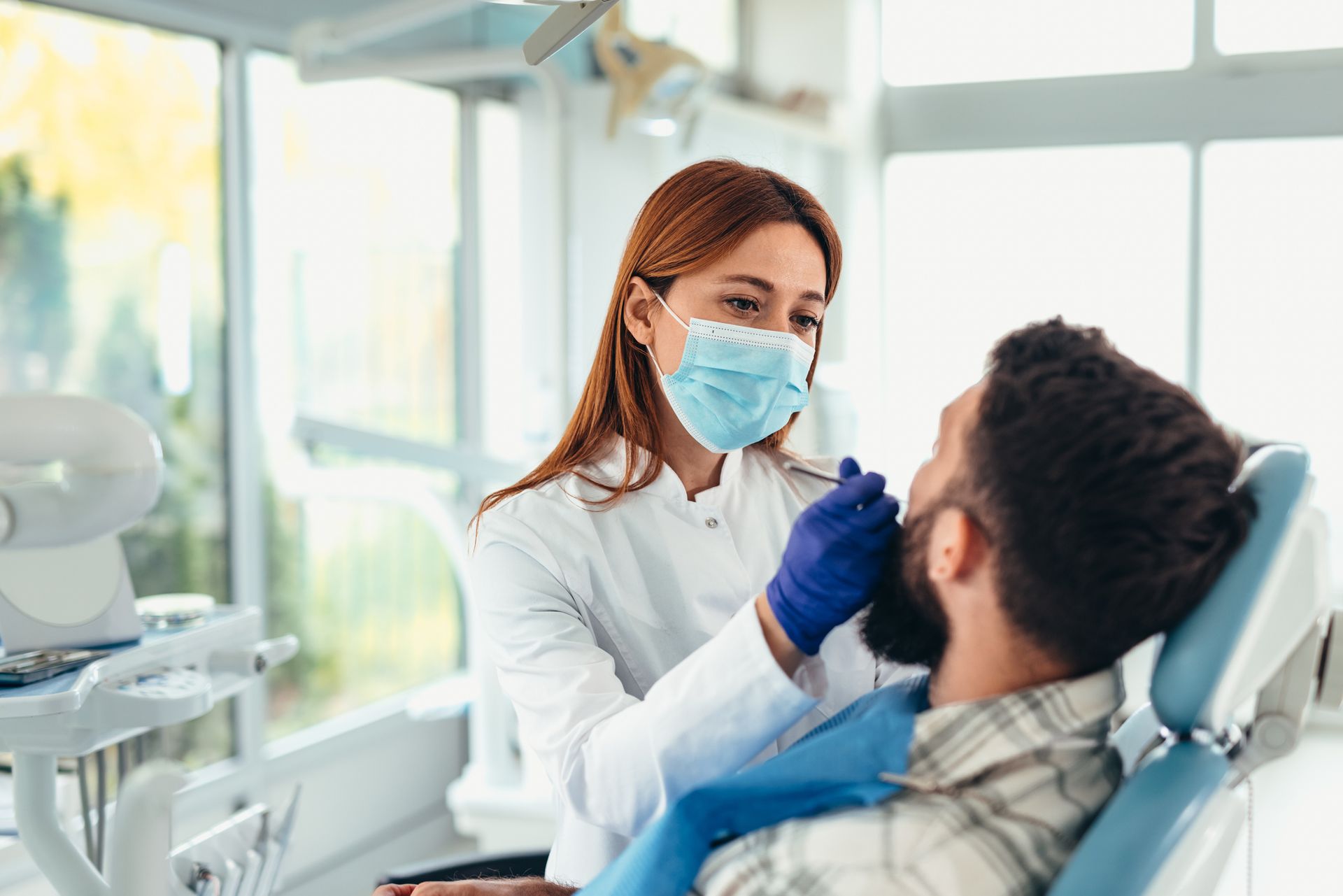 Dentist examining patient's teeth with tools in a dental office. The dentist wears a mask and gloves.