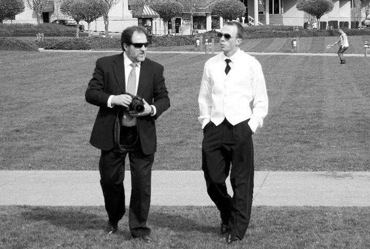 Bride and groom dance outdoors on grass; man in vest, woman in white gown, and guest in background.