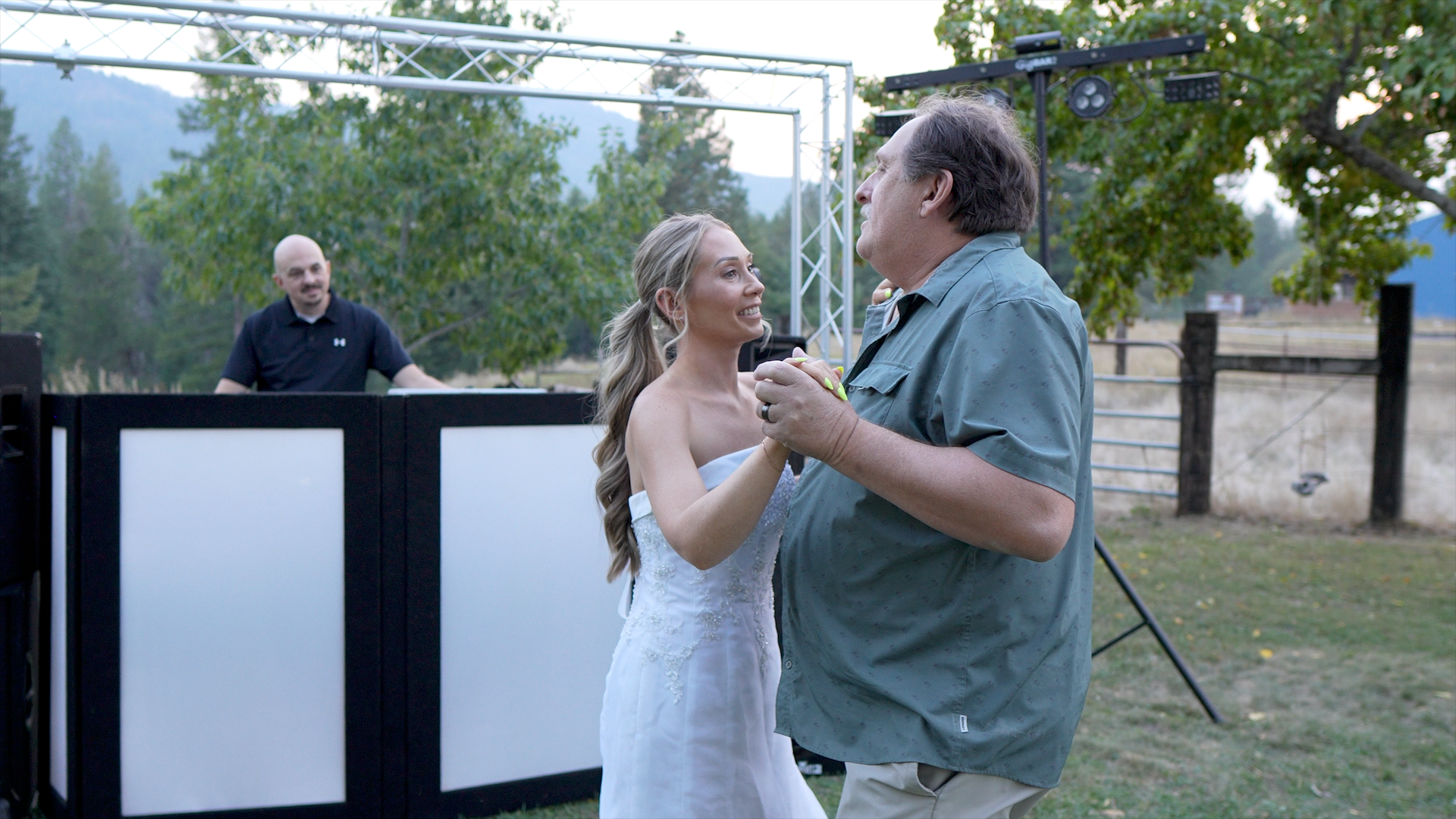 A bride dances with a man outdoors near a DJ setup, mountains in the background.