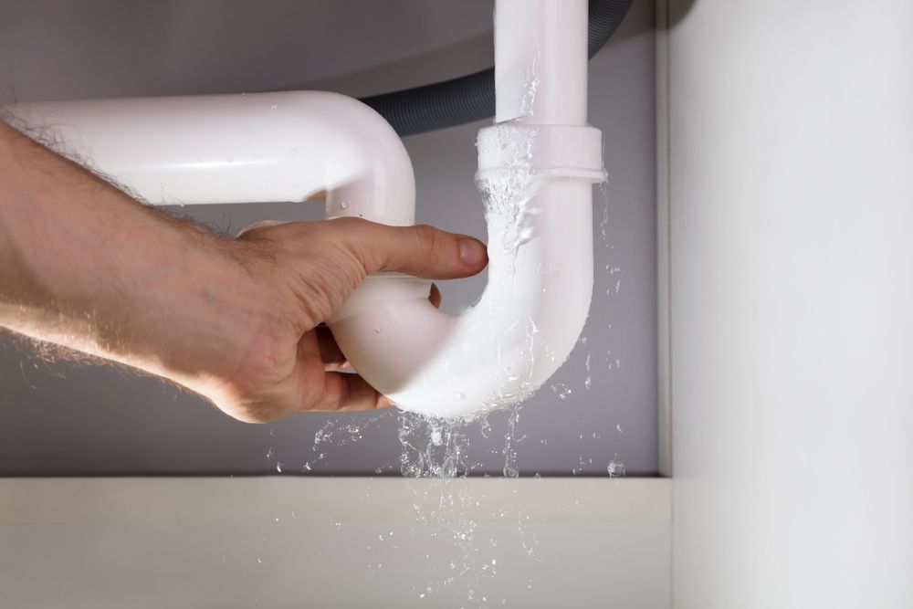 Hand Holding a Leaking White Pipe Under a Sink. Water is Spraying — Pure Plumbing & Electrical in Orange, NSW