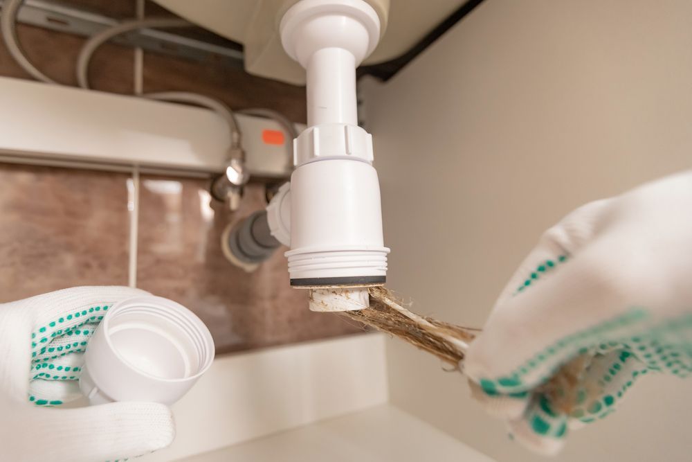 Hands in Gloves Unscrew a Sink Drain Trap Under a Cabinet — Pure Plumbing & Electrical in Parkes, NSW