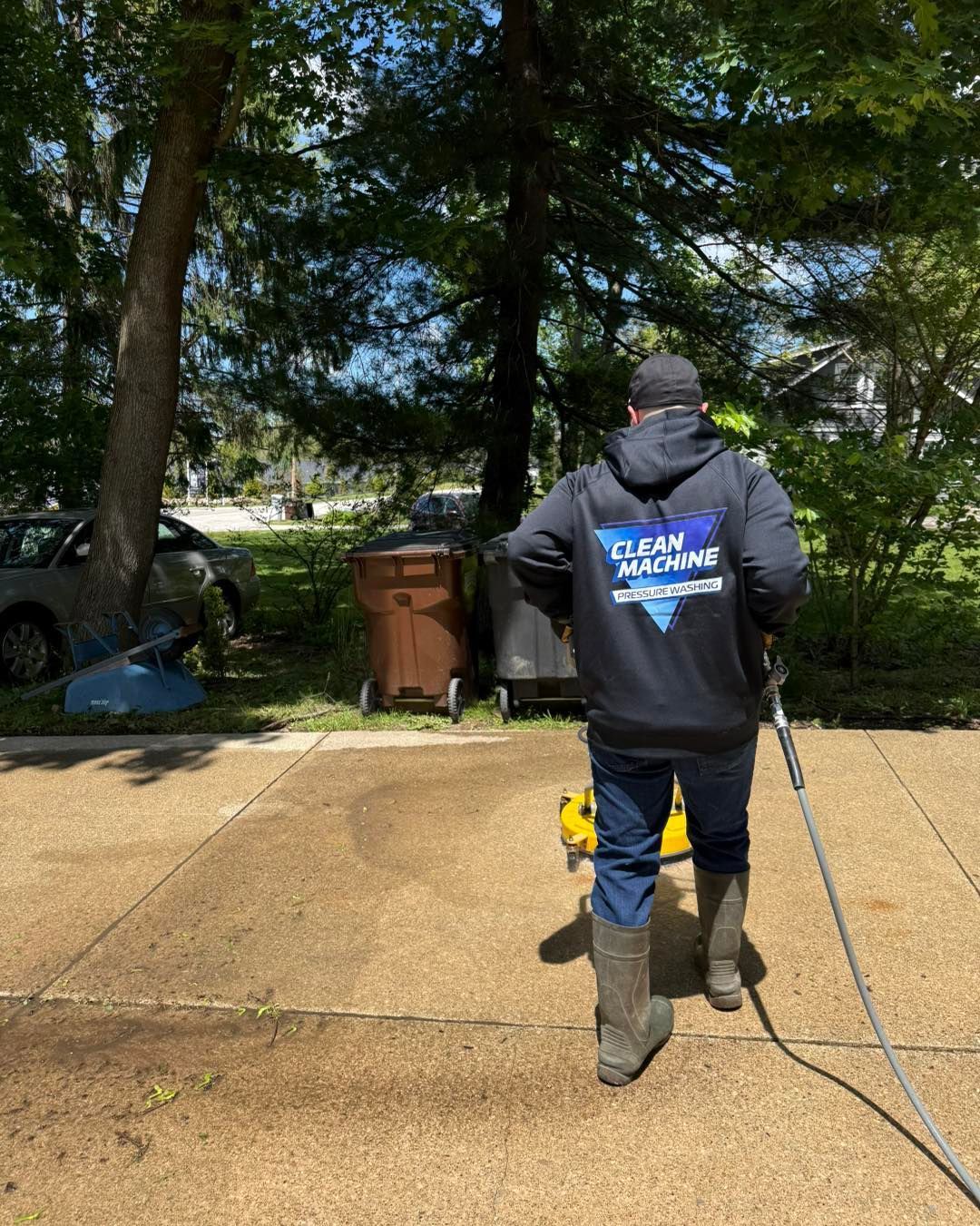 Worker in blue hoodie spraying a driveway with a pressure washer beside trash bins and trees.
