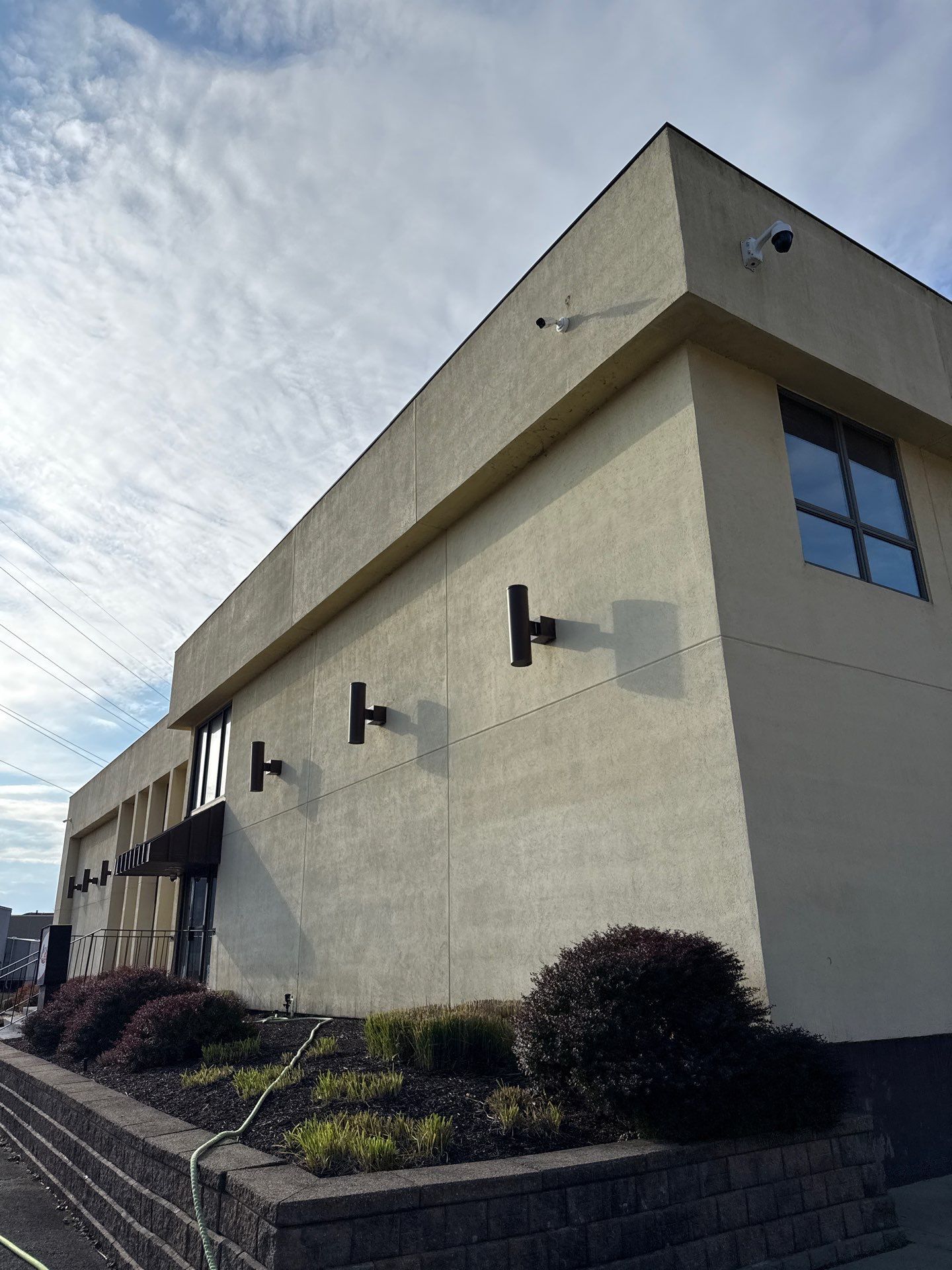 dirty commercial building with tan stucco walls, wall lights, and landscaped shrubs under a cloudy sky