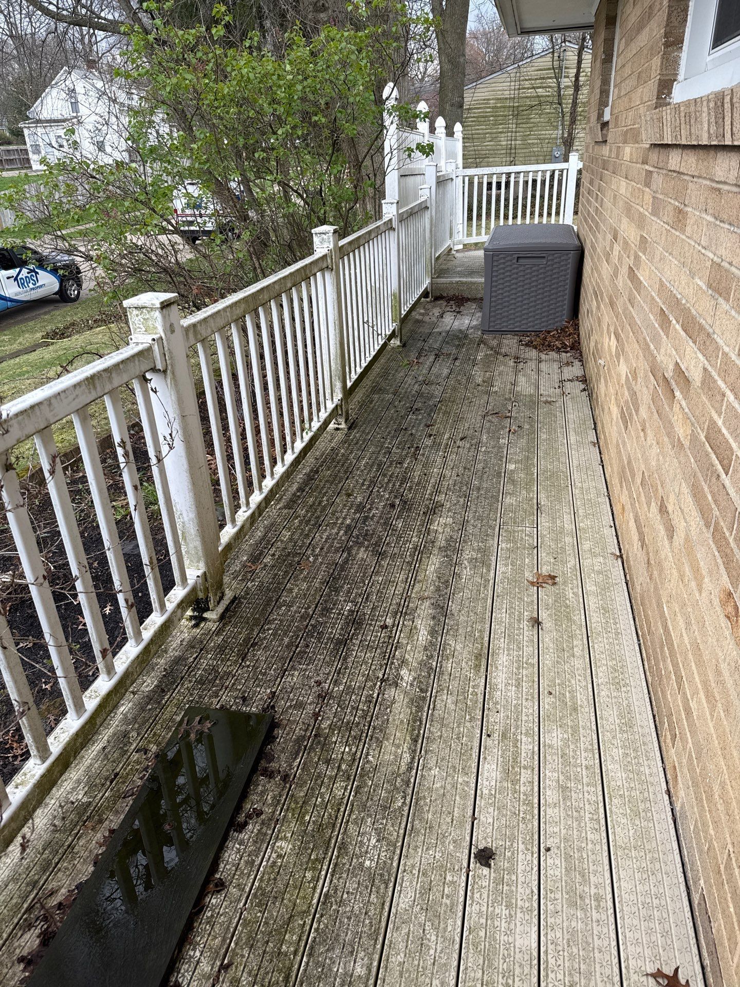Narrow wooden balcony deck with white railing, weathered boards, and a gray storage box beside a brick wall