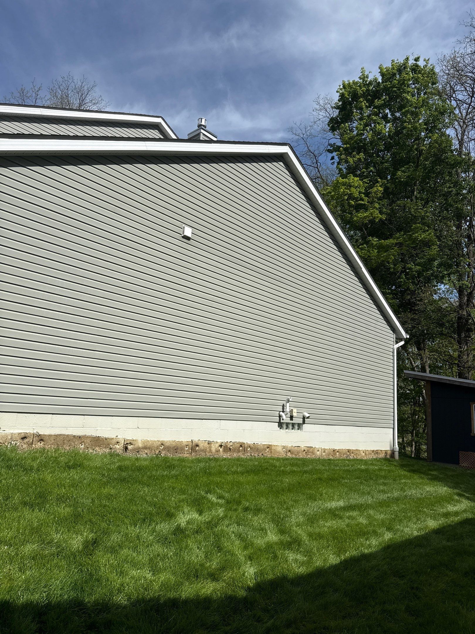 Gray house side with wavy siding, green lawn, and trees under a cloudy sky