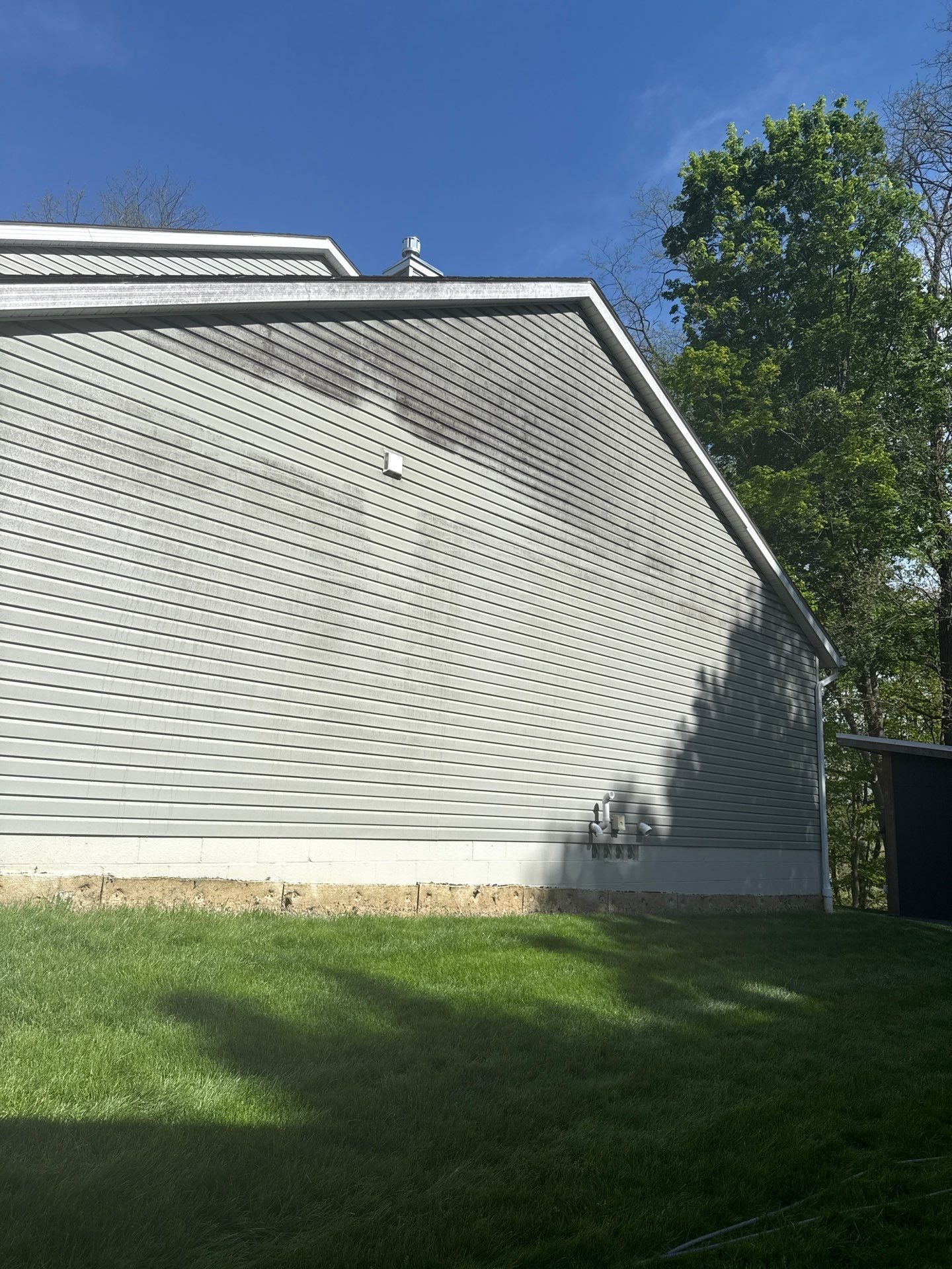 Gray house side with white trim, grassy yard, and a tall tree under a clear blue sky