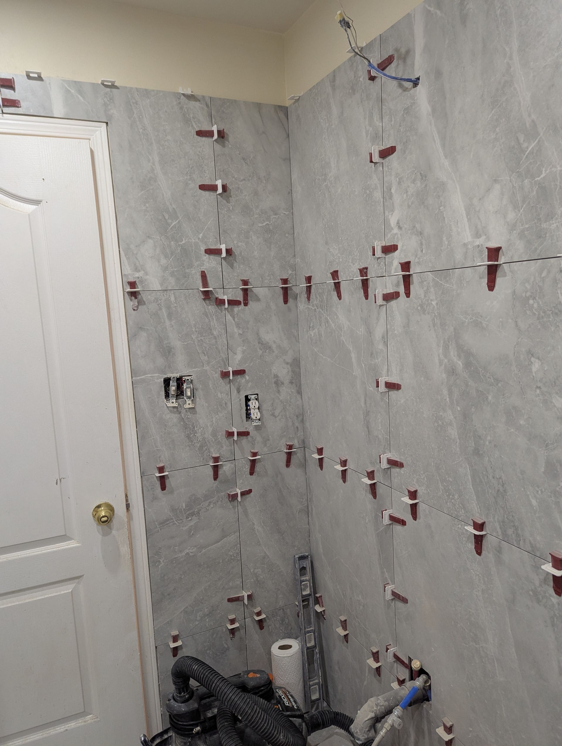 Gray tiled bathroom walls with leveling clips; door on left, electrical outlets visible.