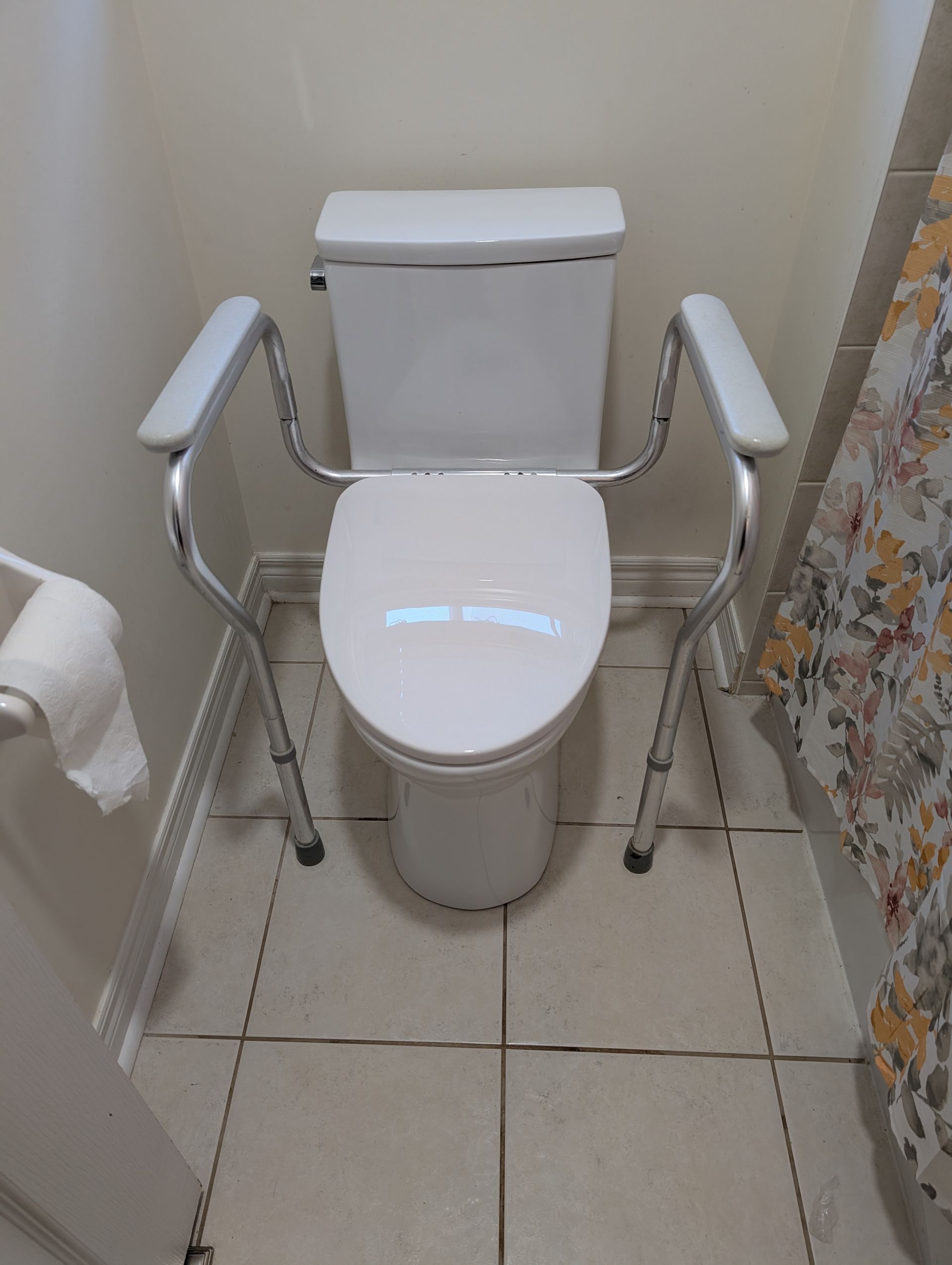 Toilet with raised seat and safety arms in a small bathroom. Beige tiled floor, white toilet.