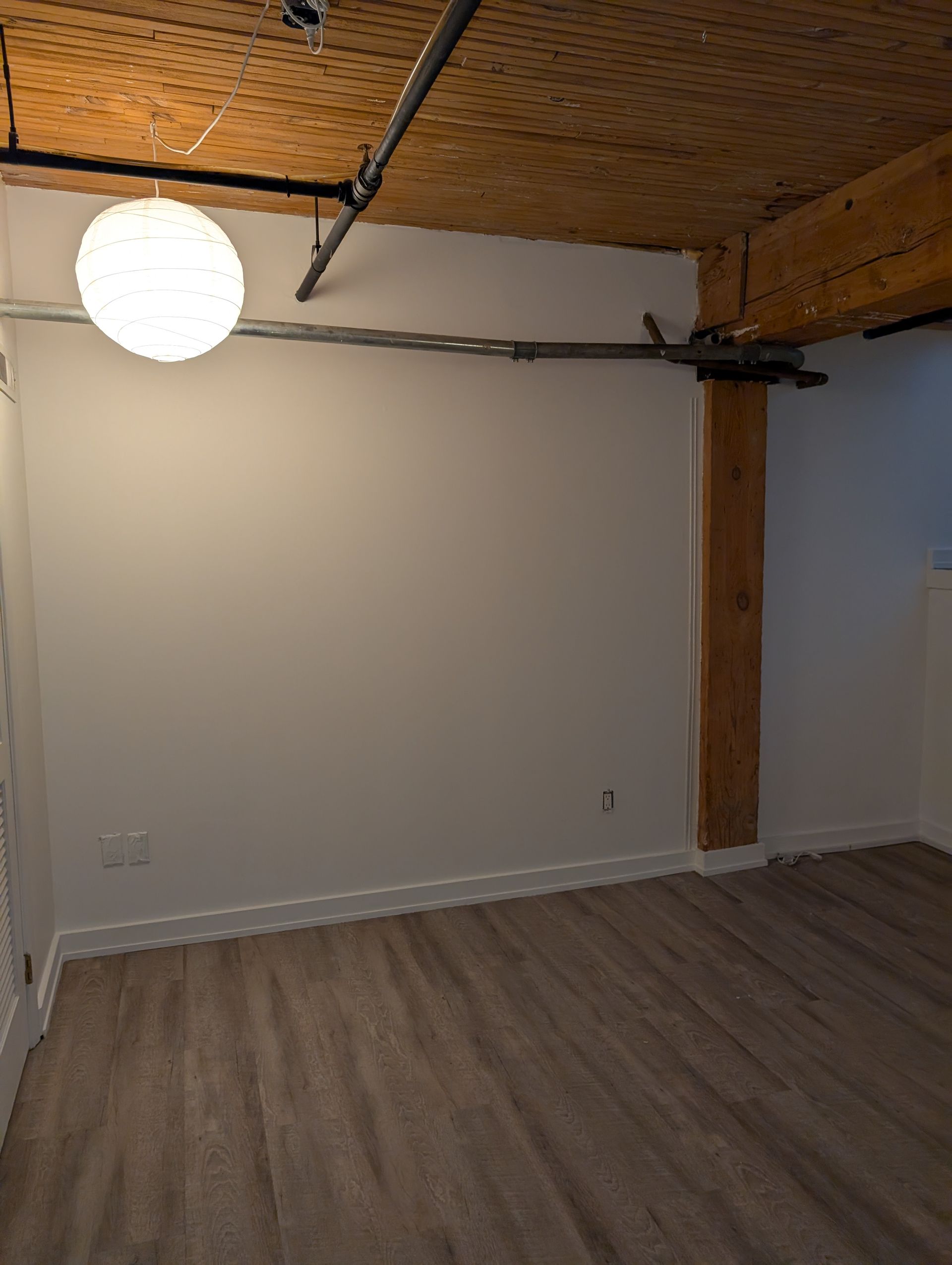 Empty room with white walls, exposed wooden beam, and wood-look flooring. A globe-shaped light hangs from the ceiling.
