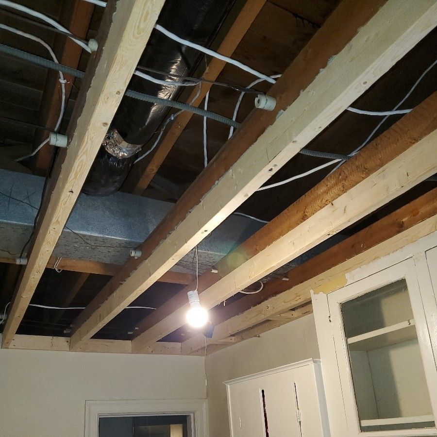 Exposed ceiling beams with wiring and ductwork, a lightbulb hangs down. Cabinets and a doorframe are visible.