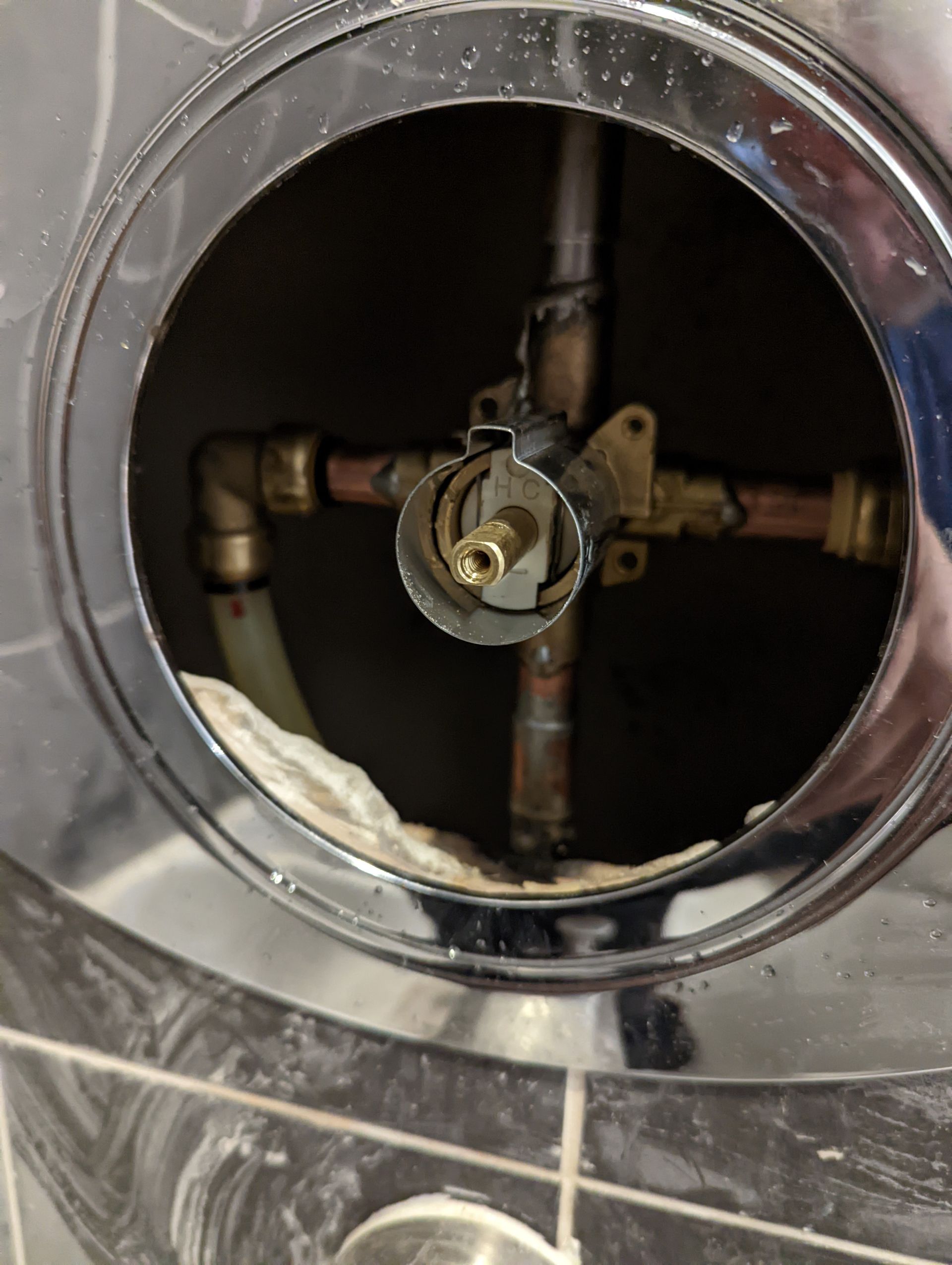 Close-up of a shower valve with plumbing connections visible through an opening. Chrome trim surrounds the opening.