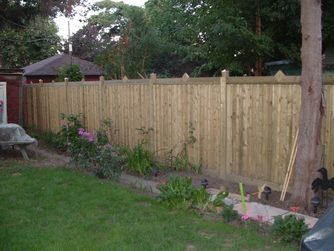 Wooden fence in a backyard, bordering a flowerbed and a lawn, with trees in the background.