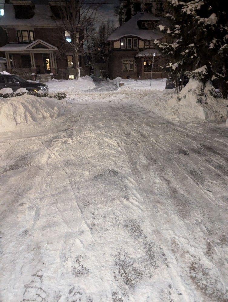 Snow-covered street with houses in the background; icy conditions, possibly slippery for pedestrians.