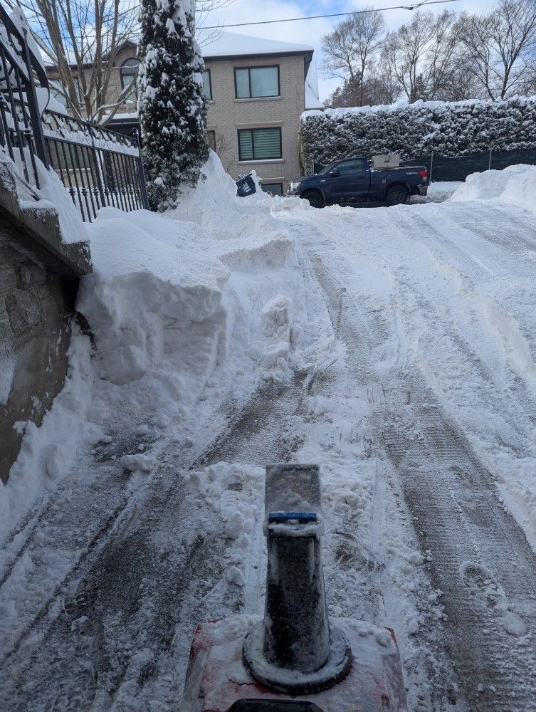 A snow-covered driveway with a snowblower clearing a path, blue truck in the background.