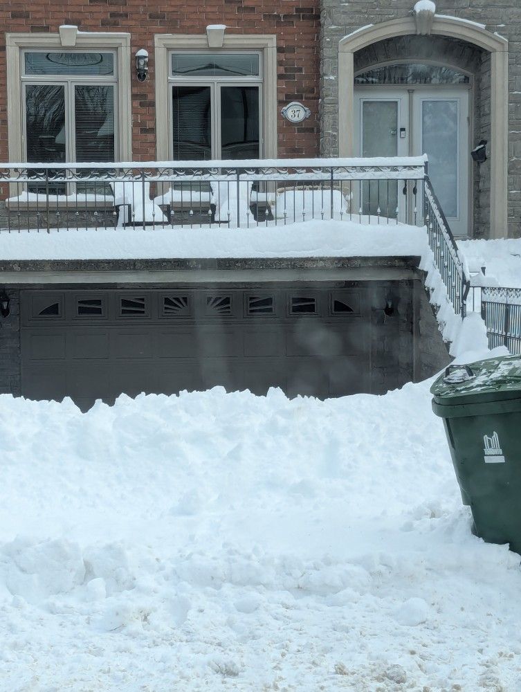 Snow-covered house with a garage. Snow piled high. Green trash can in front.