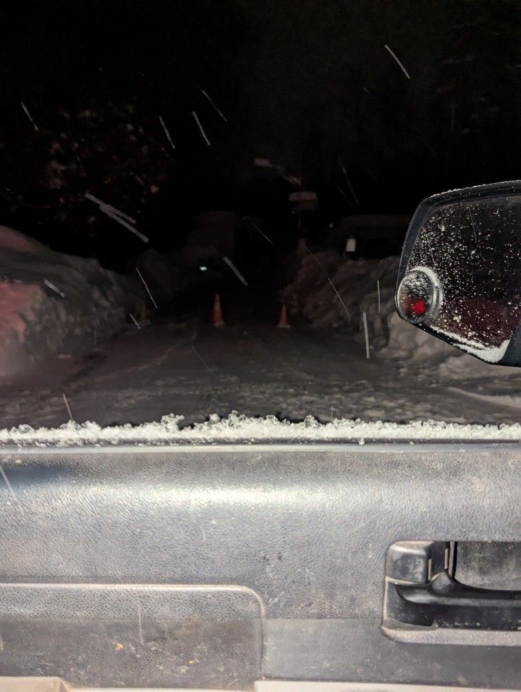 Snowy road at night; car tail light on right, snowbanks line sides, orange cones in distance.