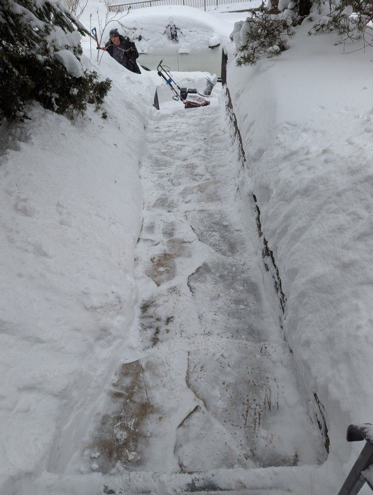 Person using a snowblower on cleared steps, surrounded by snowbanks. Snowy outdoor setting.