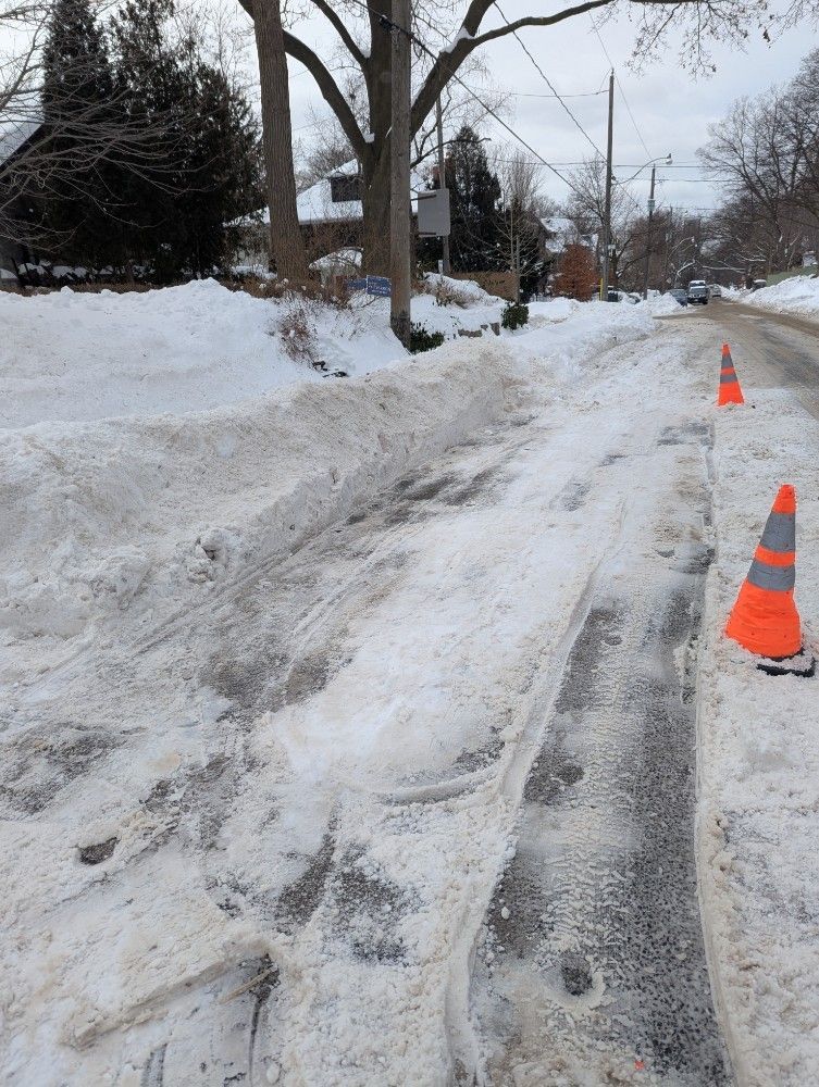 Snow-covered road with deep tire tracks and orange cones. Residential area, winter scene.