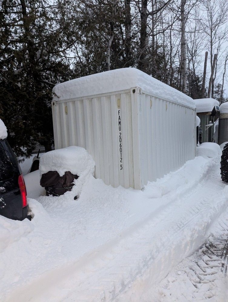 White storage container in snow, surrounded by tire tracks and trees.