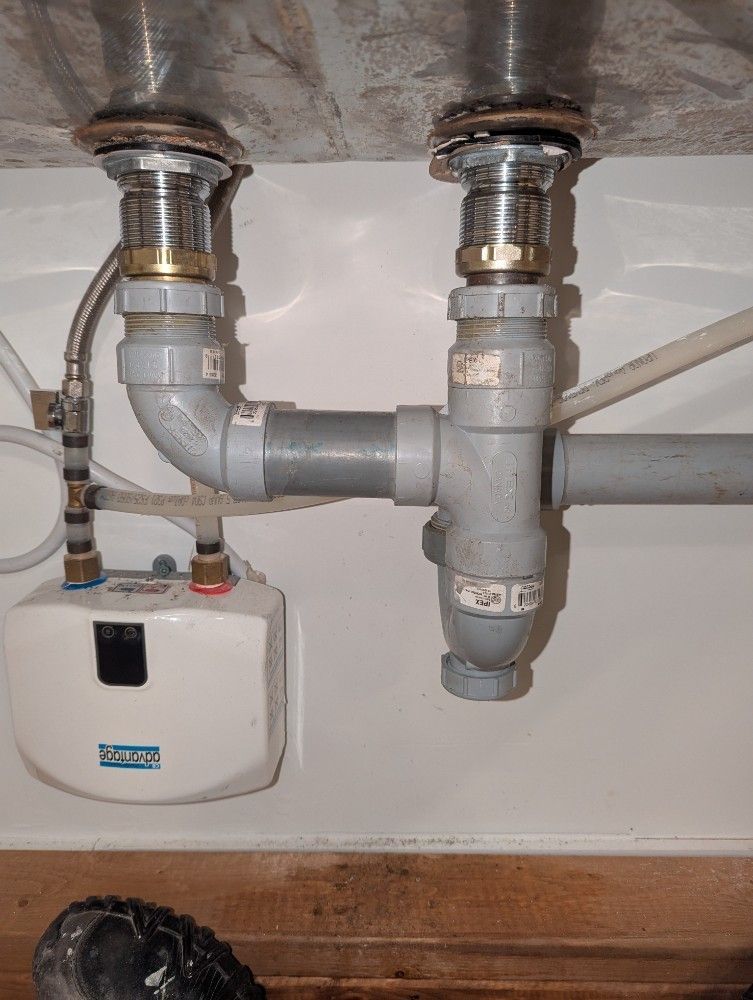 Plumbing under a sink with drains, pipes, and a small water heater. White, gray, and metal components.